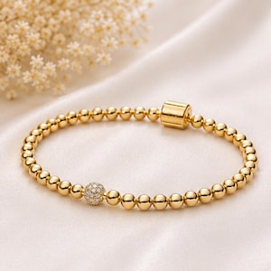 May include: Gold-colored beaded bracelet with a small, round, diamond-like accent. The bracelet is made of small, evenly spaced beads and has a cylindrical clasp. The background is a soft, white fabric.