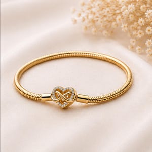 Könnte beinhalten: Goldfarbenes Armband mit herzförmigem Verschluss. Der Verschluss zeigt ein Unendlichkeitssymbol-Design, das mit kleinen, klaren Edelsteinen besetzt ist. Das Armband hat eine strukturierte Schlangenketten-Optik.
