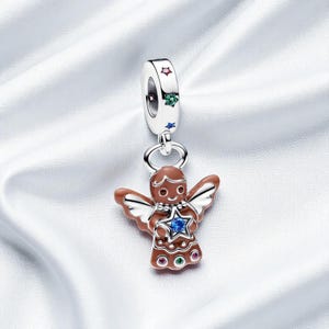 Puede incluir: Un charm de plata con un colgante de ángel de jengibre. El ángel es marrón con alas blancas y una estrella azul. El charm tiene detalles de estrellas de colores.