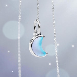 Può includere: Un ciondolo a forma di mezzaluna argentata con una superficie iridescente blu chiaro e bianca è appeso a una delicata catena. Il ciondolo ha una finitura lucida ed è sospeso su uno sfondo morbido e sfocato con accenti scintillanti.