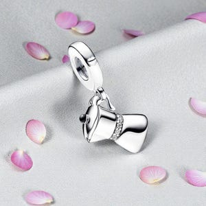 Könnte beinhalten: Ein silberner Charm-Anhänger in Form einer Kaffeemaschine, mit einer Schlaufe für ein Armband oder eine Halskette. Der Charm hat eine Reihe kleiner, klarer Steine am Boden und einen schwarzen Akzent oben. Rosa Blütenblätter sind um den Charm verstreut.
