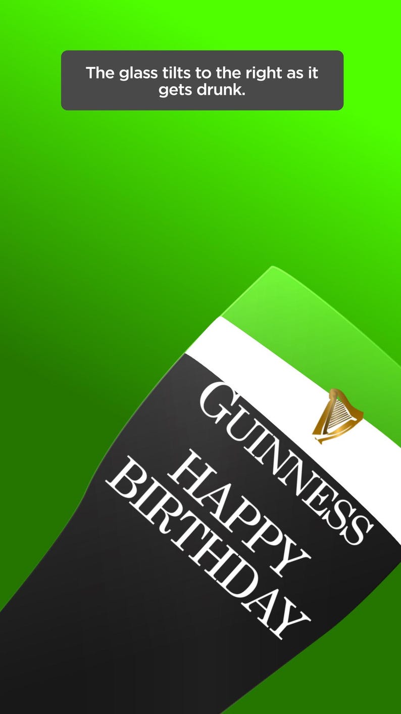 May include: A tilted Guinness Happy Birthday glass against a green background. The glass has a black section with white text and a gold harp logo. A text box at the top says, "The glass tilts to the right as it gets drunk."