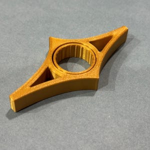 May include: A gold-colored, star-shaped object with a central circular opening and triangular cutouts. The object appears to be 3D-printed and sits on a gray surface. The design is symmetrical and geometric.