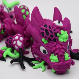 May include: A close-up of a purple dragon sculpture with green and black accents. The dragon has large white eyes with black pupils, and green and black details on its head and body. The sculpture is on a white background.