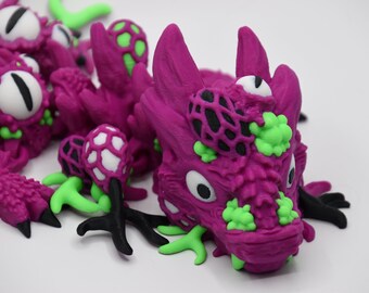 May include: A close-up of a purple dragon sculpture with green and black accents. The dragon has large white eyes with black pupils, and green and black details on its head and body. The sculpture is on a white background.