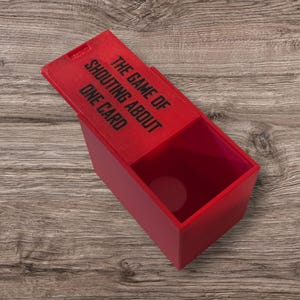 May include: A red rectangular box with a lid, featuring the text "THE GAME OF SHOUTING ABOUT ONE CARD" in black. The lid is slightly ajar, revealing the interior of the box. The box is on a wooden surface.