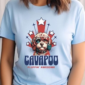 Könnte beinhalten: Ein hellblaues T-Shirt mit einem Cartoon-Bild eines Cavappoo-Hundes, der eine Sonnenbrille trägt. Der Hund ist von roten, weißen und blauen Sternen umgeben. Der Text "CAVAPOO" ist in einer Retro-Schrift unter dem Hund geschrieben, und der Text "FLUFFIN' AWESOME" ist darunter geschrieben.