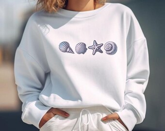 Könnte beinhalten: Weißer Pullover mit einem Aufdruck von Muscheln und einem Seestern. Der Aufdruck ist in Blautönen und Lila.