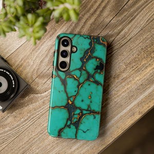 May include: A turquoise phone case with a gold and black veined pattern. The case is on a wooden surface, with a camera and plant in the background. The case has a three-lens camera cutout.
