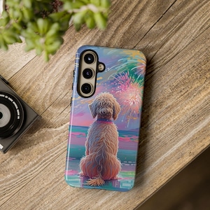 Könnte beinhalten: Eine Handyhülle mit einem Hund, der sich ein Feuerwerk über dem Meer ansieht. Die Hülle hat ein farbenfrohes Design mit einem Hund im Vordergrund und Feuerwerk im Hintergrund. Die Hülle liegt auf einer Holzoberfläche.