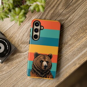 May include: A colorful phone case with a bear illustration. The case features horizontal stripes in red, teal, yellow, and orange. The bear is brown with detailed fur and a focused expression. The case is on a wooden surface.