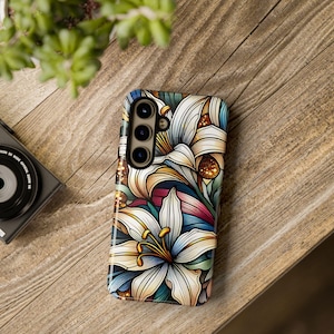 Puede incluir: Una funda para teléfono con un diseño floral colorido que presenta lirios blancos, detalles dorados y detalles azules y rojos.
