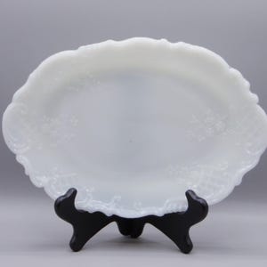 May include: White milk glass oval serving dish with a raised floral design. The dish is on a black stand.