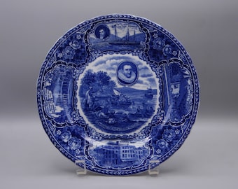 Puede incluir: Plato de cerámica azul y blanco con un diseño decorativo que presenta figuras históricas y escenas de la fundación de la ciudad de Nueva York. El plato incluye imágenes de Peter Minuit, el desembarco de Hendrick Hudson y los primeros edificios de la ciudad.