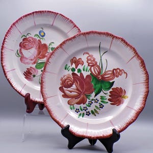 May include: Two white ceramic plates with pink and red floral designs. The plates have a scalloped edge and a pink rim.