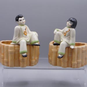 May include: A pair of ceramic planters shaped like bamboo stalks with a seated figurine of a person on each. The figurines are wearing traditional Asian clothing and have black hair.