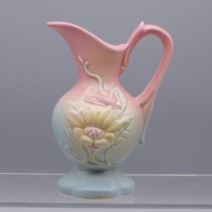 May include: A pastel-colored ceramic pitcher with a floral design. The pitcher features a light pink handle and spout, with a large, embossed yellow flower on the front. The base is a light blue color, and the overall design is delicate and vintage.