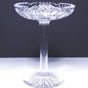 May include: A clear, cut-glass pedestal bowl with a scalloped rim. The bowl sits atop a slender stem and a circular base, showcasing intricate geometric patterns. The design reflects light, creating a sparkling effect, ideal for serving desserts or displaying decorative items.