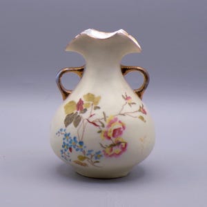 May include: A small, white porcelain vase with gold accents and a floral design. The vase has two handles and a ruffled top.