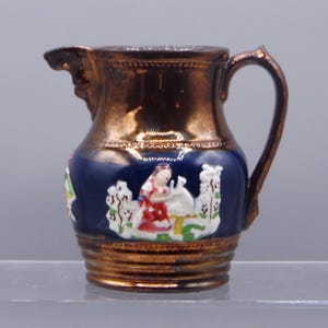 May include: A small, antique copper-colored pitcher with a dark blue band featuring a painted scene of a person and a swan. The pitcher has a handle and a spout. The design is detailed and the colors are vibrant.