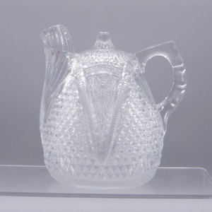 May include: A clear glass pitcher with a decorative handle and spout. The pitcher features a diamond-cut pattern across its surface, with a central design of vertical lines and a starburst. The pitcher is likely used for serving beverages.