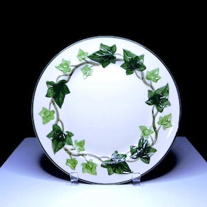 May include: White ceramic plate with a green ivy leaf design around the rim. The leaves are in varying shades of green, with a dark green border. The plate is displayed on a clear stand.