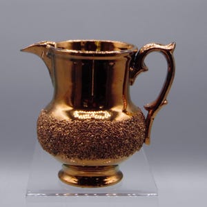 May include: A small copper-colored ceramic pitcher with a textured base and a handle. The pitcher has a shiny, metallic finish.