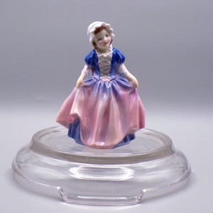 May include: A porcelain figurine of a girl wearing a blue and pink dress with a white bonnet. She is standing on a clear glass base.