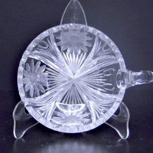 May include: Clear, cut-glass bowl with a handle, resting on a clear stand. The bowl features a floral and starburst design. The handle is small and rounded. The bowl is likely for serving or display.