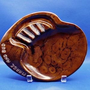 May include: A brown ceramic ashtray with a glossy finish and a mottled, dark brown pattern. The ashtray features four integrated cigarette rests and the embossed text MARGE AND BUD along the curved edge of the rounded, irregular shape.