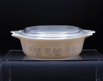 May include: A tan Pyrex casserole dish with a clear glass lid. The dish features a silver ivy leaf pattern around the base. The dish has two handles and is sitting on a clear acrylic surface.