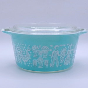 May include: A turquoise Pyrex casserole dish with a clear glass lid. The dish features a white graphic design of a rooster, people, and plants. The casserole dish has two handles.