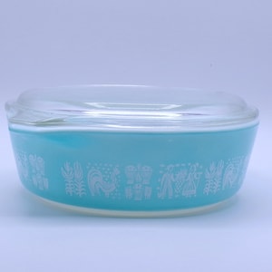May include: A vintage turquoise Pyrex casserole dish with a clear glass lid. The dish features a white, stylized pattern of figures and farm animals around the exterior. A classic piece of kitchenware.