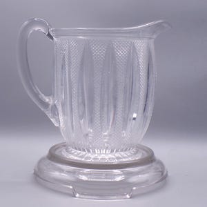 May include: A clear glass pitcher with a textured design and a round base. The pitcher has a handle and a spout.