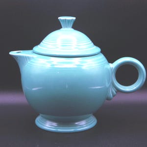 May include: A turquoise ceramic teapot with a lid and a round handle. The teapot has a spout and sits on a pedestal base. The lid has a small, decorative finial. The teapot is a solid, glossy color.