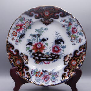 May include: A white porcelain plate with a blue and gold rim. The plate features a floral design with red, pink, and blue flowers. The center of the plate has a floral design with a black and gold border.