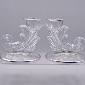 May include: A pair of clear glass candle holders with silver accents. The holders are shaped like a stylized leaf with a curled end.