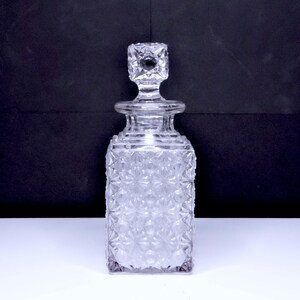 May include: A clear, square-shaped glass decanter with a geometric pattern and a square stopper. The decanter has a textured surface and is designed to hold liquids. The background is a dark color, and the decanter is sitting on a white surface.