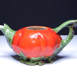 May include: A ceramic creamer shaped like a tomato. The creamer is primarily red with green accents on the handle, spout, and base. The handle is curved, and the spout is pointed. The creamer is designed to resemble a tomato.