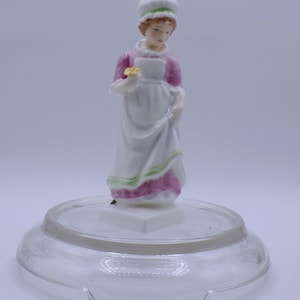 May include: A porcelain figurine of a woman in a white dress with pink and green accents, holding a yellow flower. The figurine is on a clear glass base.