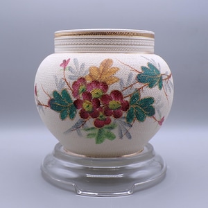 May include: Cream-colored ceramic vase with a floral design. The vase features a textured surface and is adorned with colorful flowers and leaves in shades of red, green, and gold. The rim is decorated with a gold band.