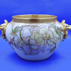May include: A white porcelain bowl featuring a hand-painted floral design with soft yellow, green, and purple hues outlined in gold. The bowl has a wide gold-toned rim and two decorative handles shaped like lion heads with a metallic gold finish.