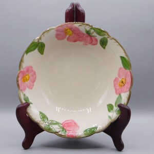 May include: A white ceramic bowl with a scalloped rim and a floral design featuring pink roses and green leaves. The bowl is on a brown stand.