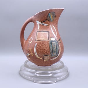 May include: A vintage brown ceramic pitcher with a curved handle and spout. The pitcher features abstract geometric designs in teal, gold, and white. The pitcher is displayed on a clear glass stand. The word "SAHARA" is visible on the side.