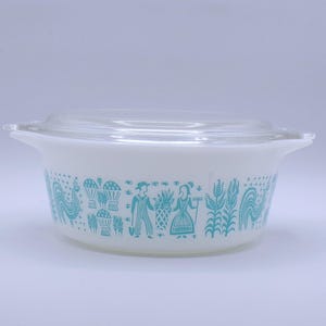 May include: White casserole dish with a clear glass lid. The dish features a turquoise design of stylized figures and plants. The dish has handles on either side and is suitable for serving food.