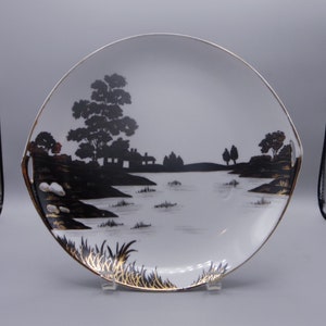 May include: White serving plate with a black landscape silhouette design featuring trees, a house, and water. The plate has a gold rim and gold accents on the landscape details. The plate has two small handles.