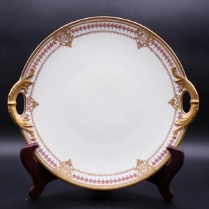 May include: A white porcelain serving platter with gold trim and handles. The rim features a delicate pink and gold floral pattern. The handles are ornate and gold. The platter is displayed on a wooden stand.
