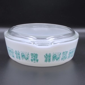 May include: White Pyrex casserole dish with a clear glass lid. The dish features a teal pattern of stylized roosters, wheat, and other designs around the rim. The dish is round with a slight handle on one side.