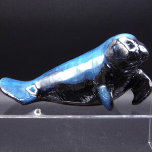 May include: A ceramic figurine of a manatee, painted in shades of blue and black. The figurine is lying on its side with its flippers outstretched.