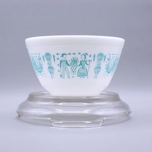 May include: White mixing bowl with a turquoise pattern featuring figures, baskets, and stylized elements. The bowl sits on a clear glass base. The design is a vintage style, suitable for kitchen decor.